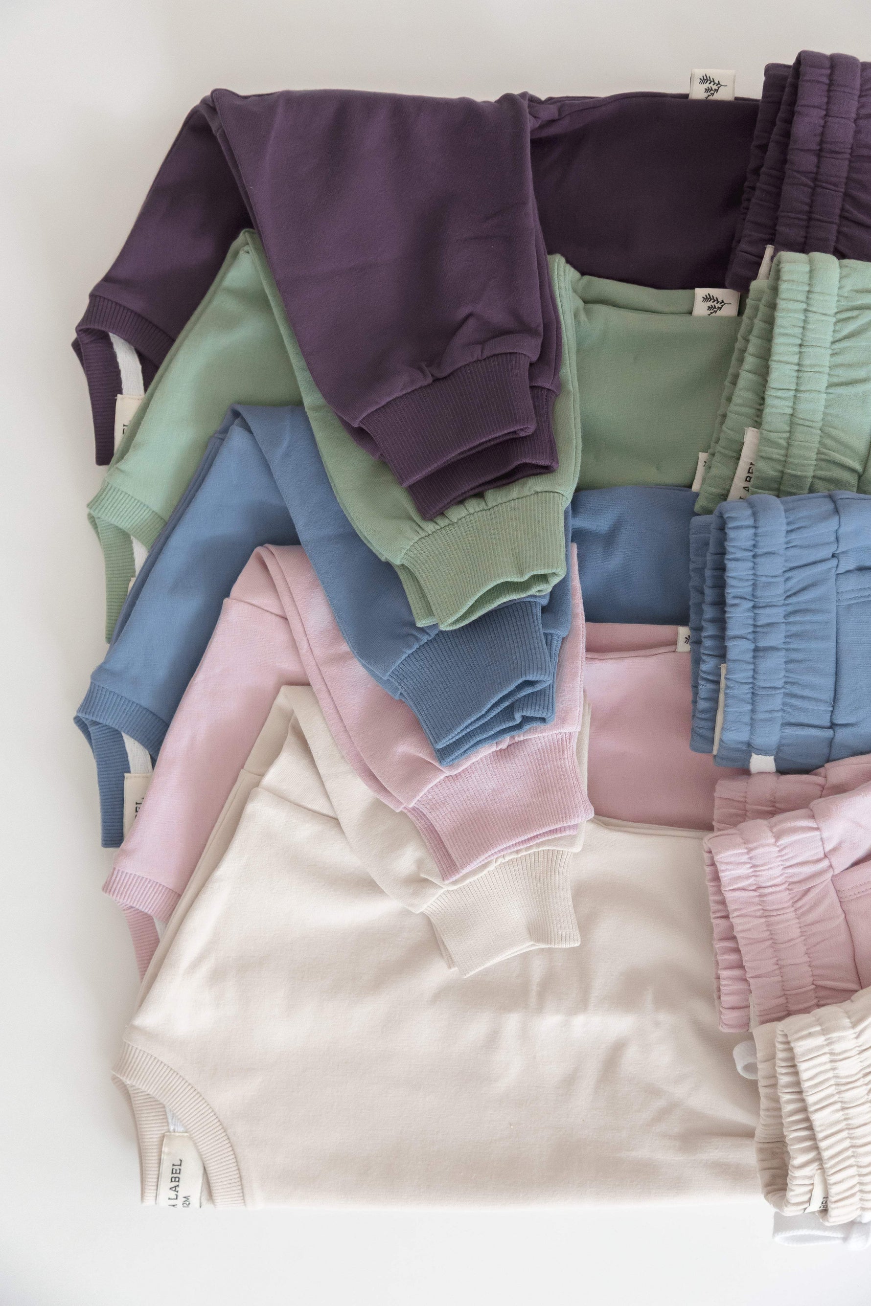 A flat-lay arrangement of folded children's loungewear in soft pastel and muted tones, including a dark purple sweatshirt, sage green sweatshirt, light blue sweatshirt, pink sweatshirt, and beige sweatshirt all on a white background.