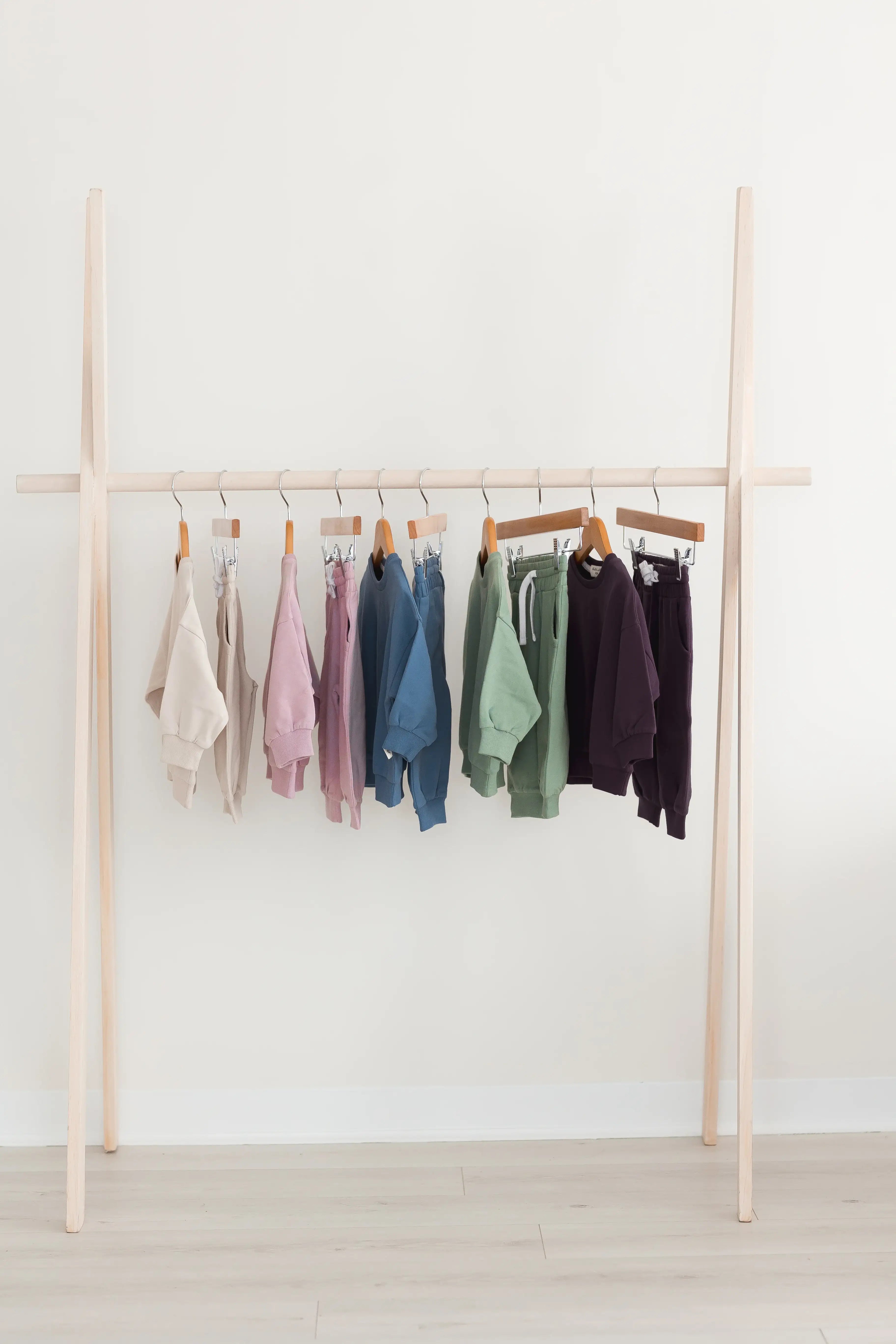 A light wood clothing rack with Milana Label's GOTS certified jogger sets hanging on wooden hangers against a plain white wall and light wood floor.