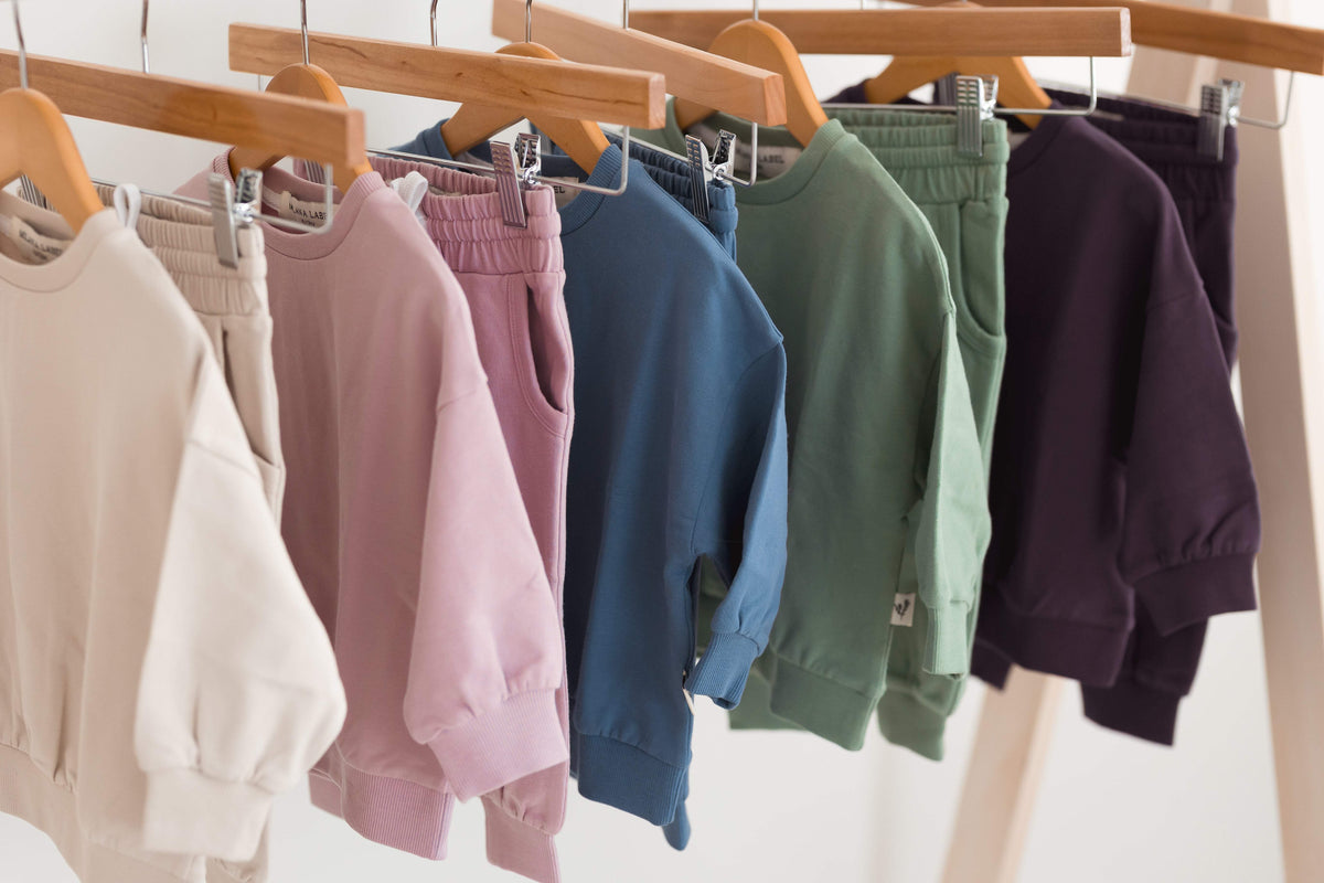 Greige, pink, blue, green and purple sweatshirts and pants for children on wooden hangers.