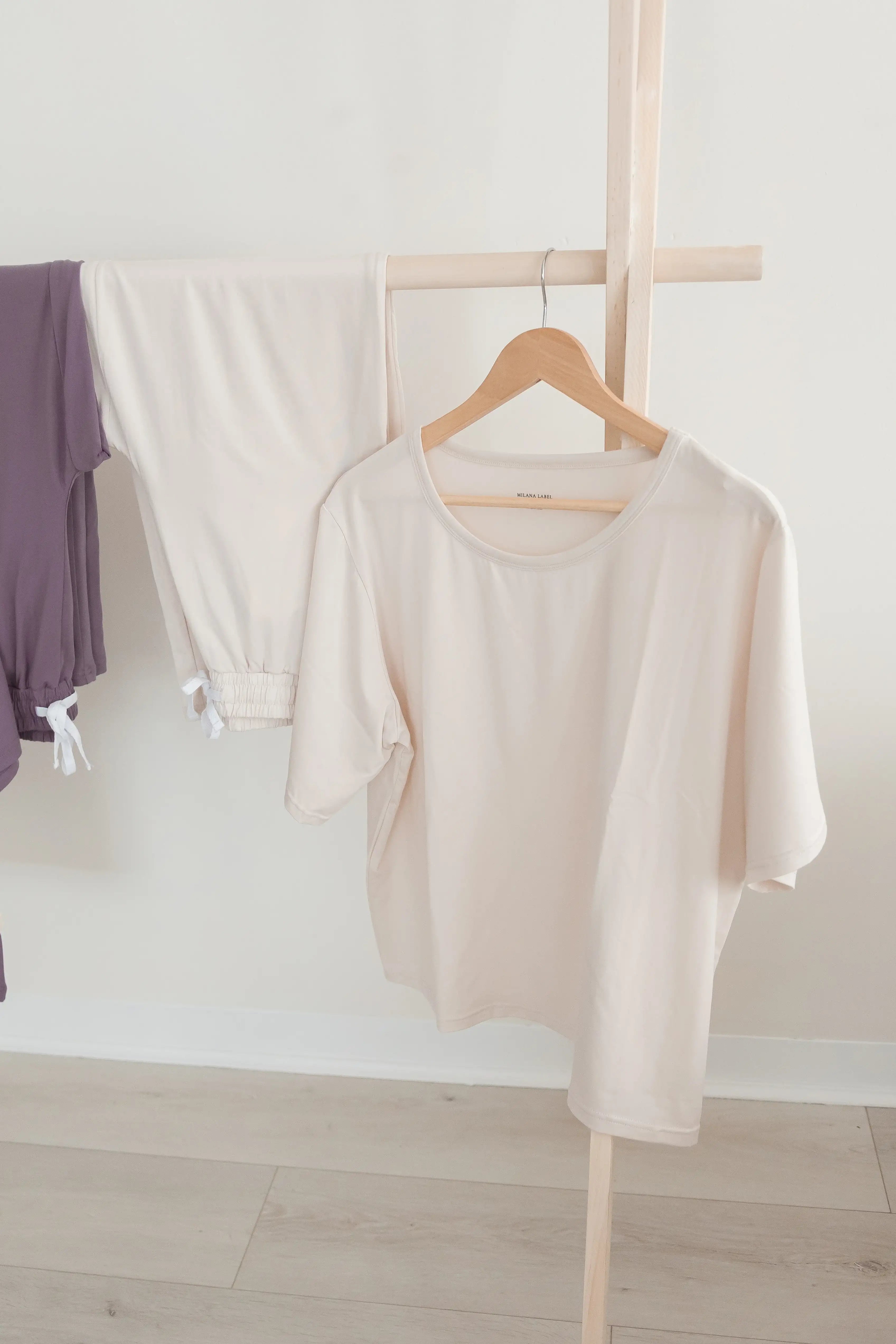 A light greige short-sleeve t-shirt hanging on a wooden hanger from a minimalist clothing rack, with matching greige pants and a purple top partially visible in the background.