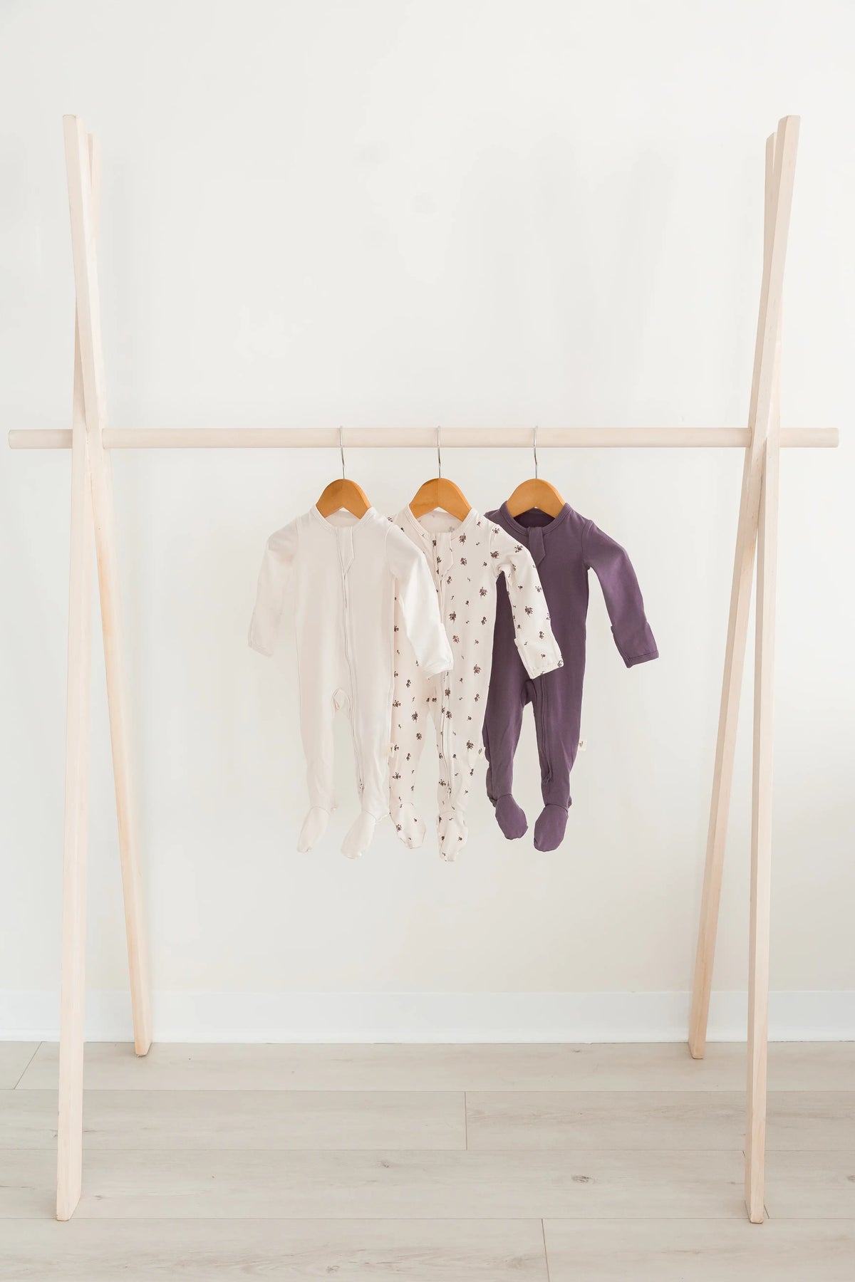 Three baby footed sleepers hanging on a light wood A-frame clothing rack against a white wall and light wood floor.