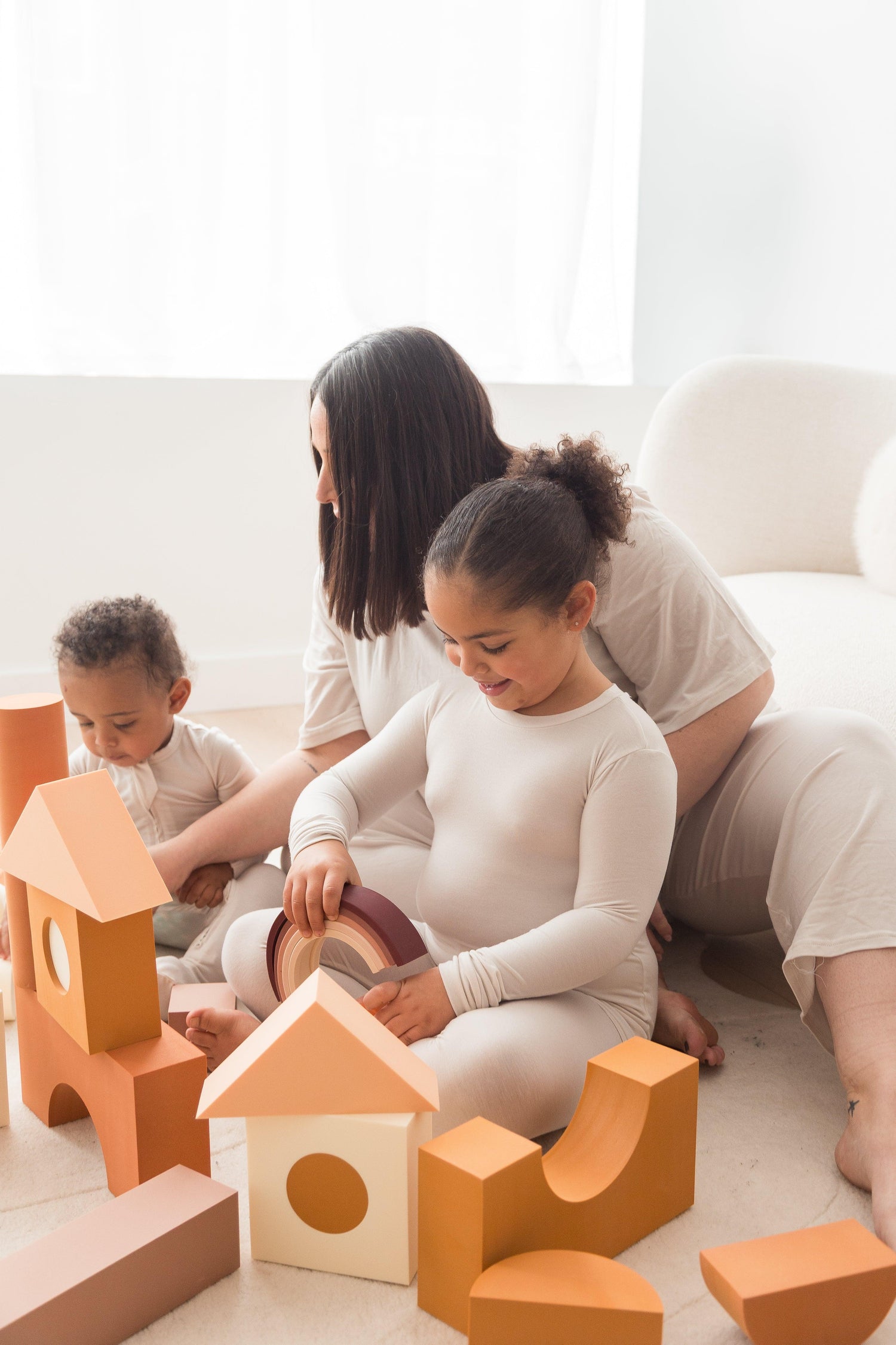 A mom and her two kids playing with blocks on the floor in their matching neutral outfits.