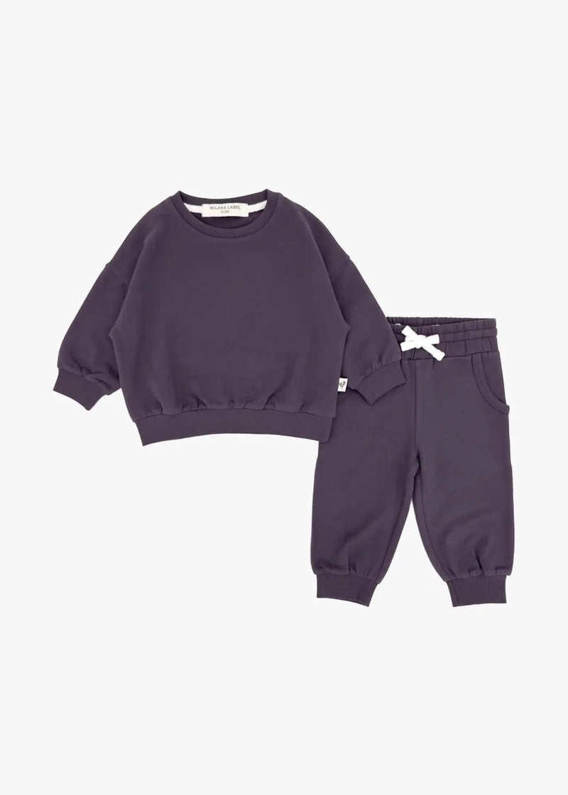 A flat lay product shot of a matching two-piece children's organic cotton loungewear set in deep elderberry purple, featuring an oversized crewneck sweatshirt and matching jogger pants against a plain white background.
