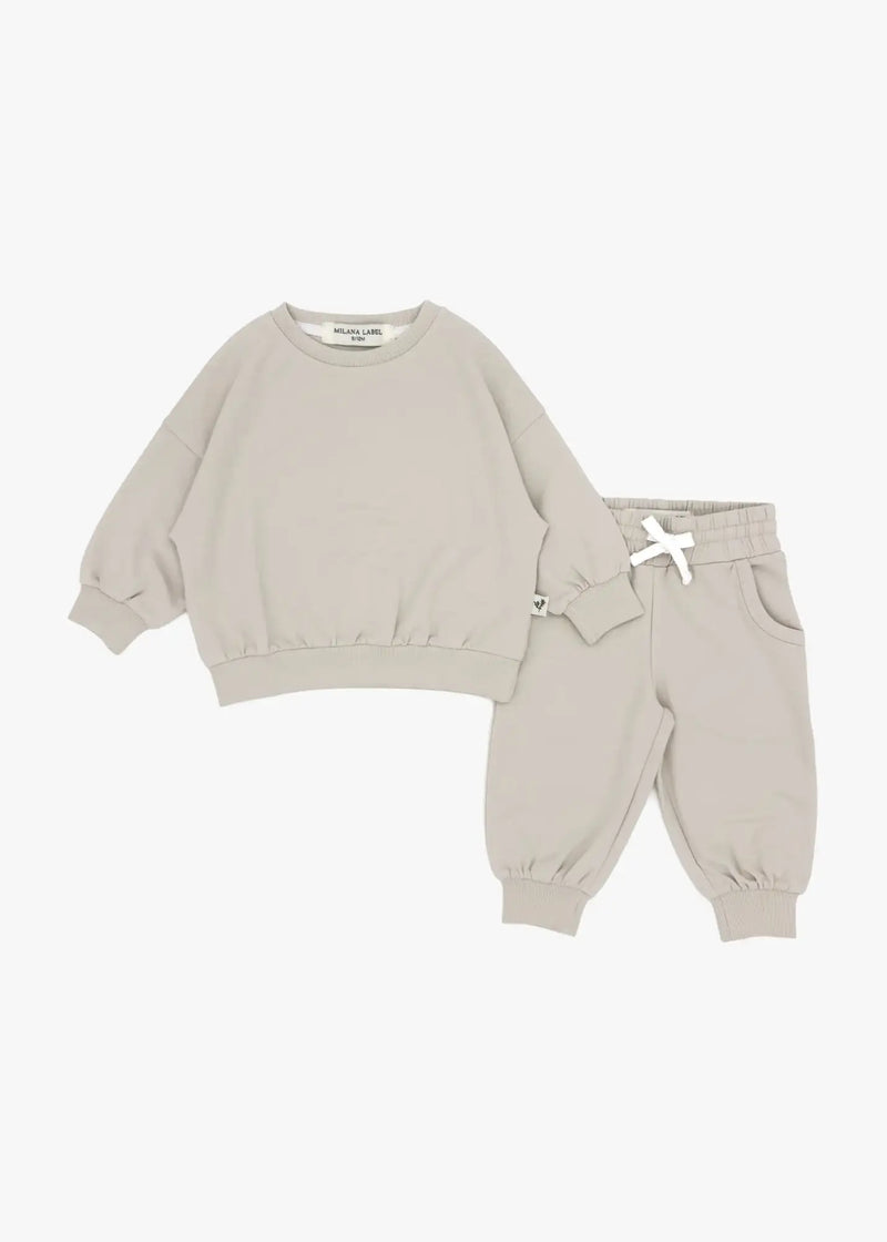 A flat lay product view of a matching two-piece children's jogger set in an oat beige color against a plain white background, featuring an oversized crewneck sweatshirt and matching jogger pants with a white drawstring bow.