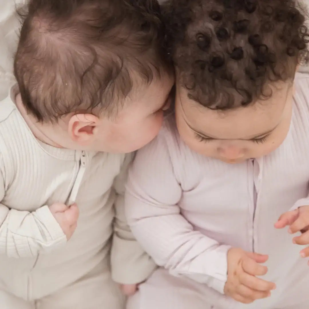 Two babies wearing matching ribbed zip-up footie onesies, one in greige and one in pale pink, lying close together on a soft neutral surface.