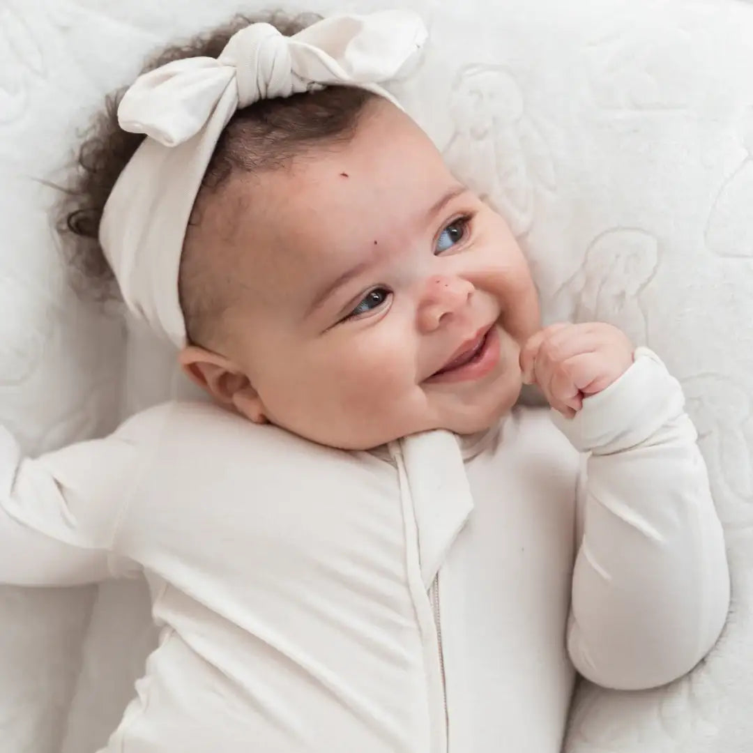 A smiling baby lying on a white quilted lounger, wearing a greige-colored long-sleeve zip-up onesie and a matching bow headband.