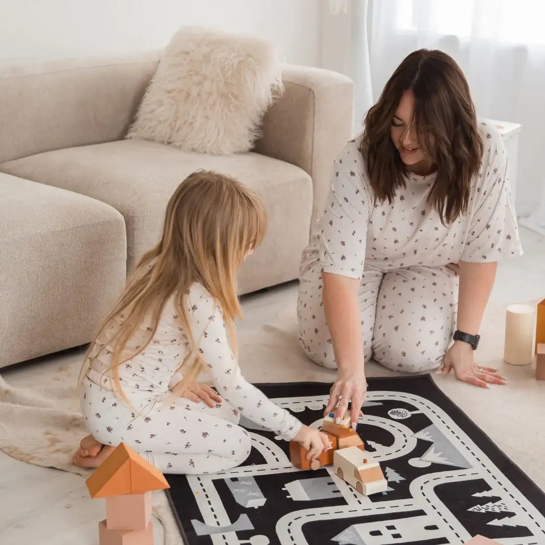 A woman and a young girl wearing matching Milana pajamas are playing on a black and white road play mat in a bright living room with a beige sofa.