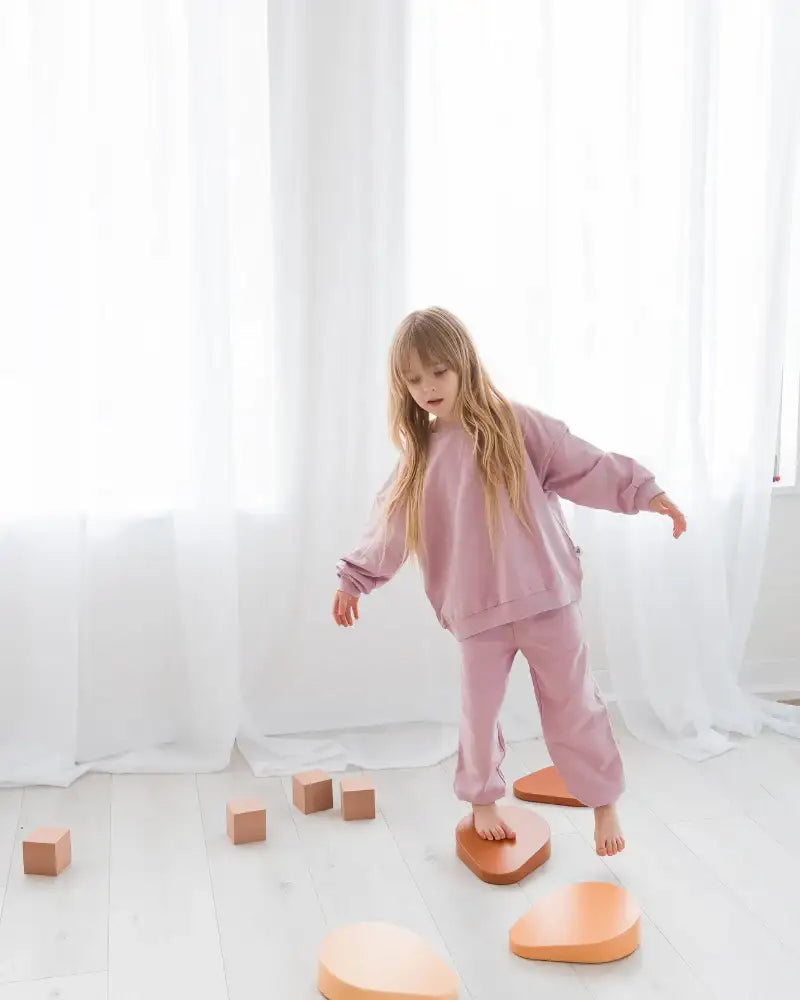 A young girl with long blonde hair is balancing on a series of peach-coloured stepping stones in a bright, minimalist room with white curtains and light wood flooring. She is wearing a matching Milana Label jogger set in the colour Pale Orchid.
