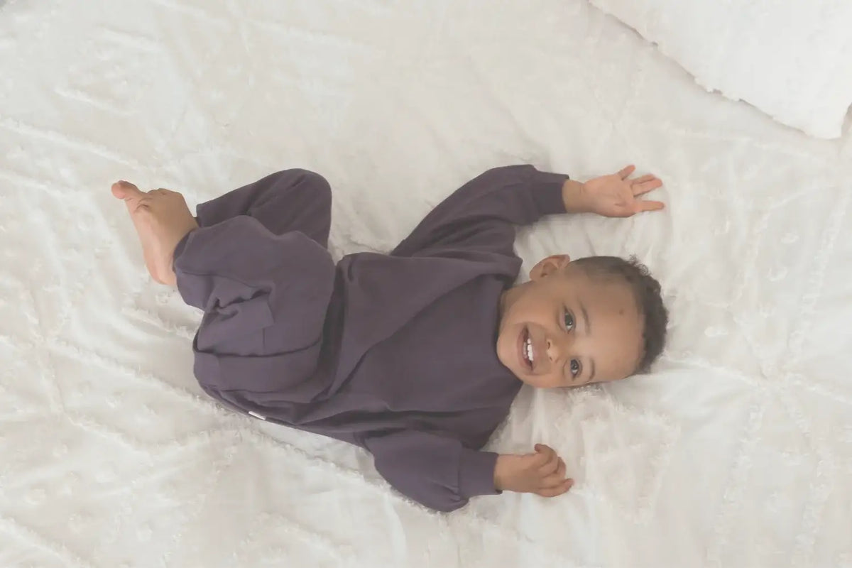 A baby wearing a purple long sleeve top and matching pants lying on a white bed.