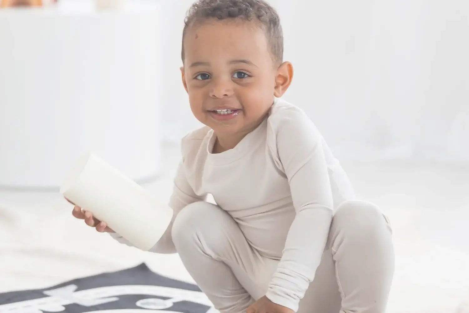 A smiling toddler wearing a light greige long-sleeve PJ set crouches on a white floor while holding a white cylindrical toy.