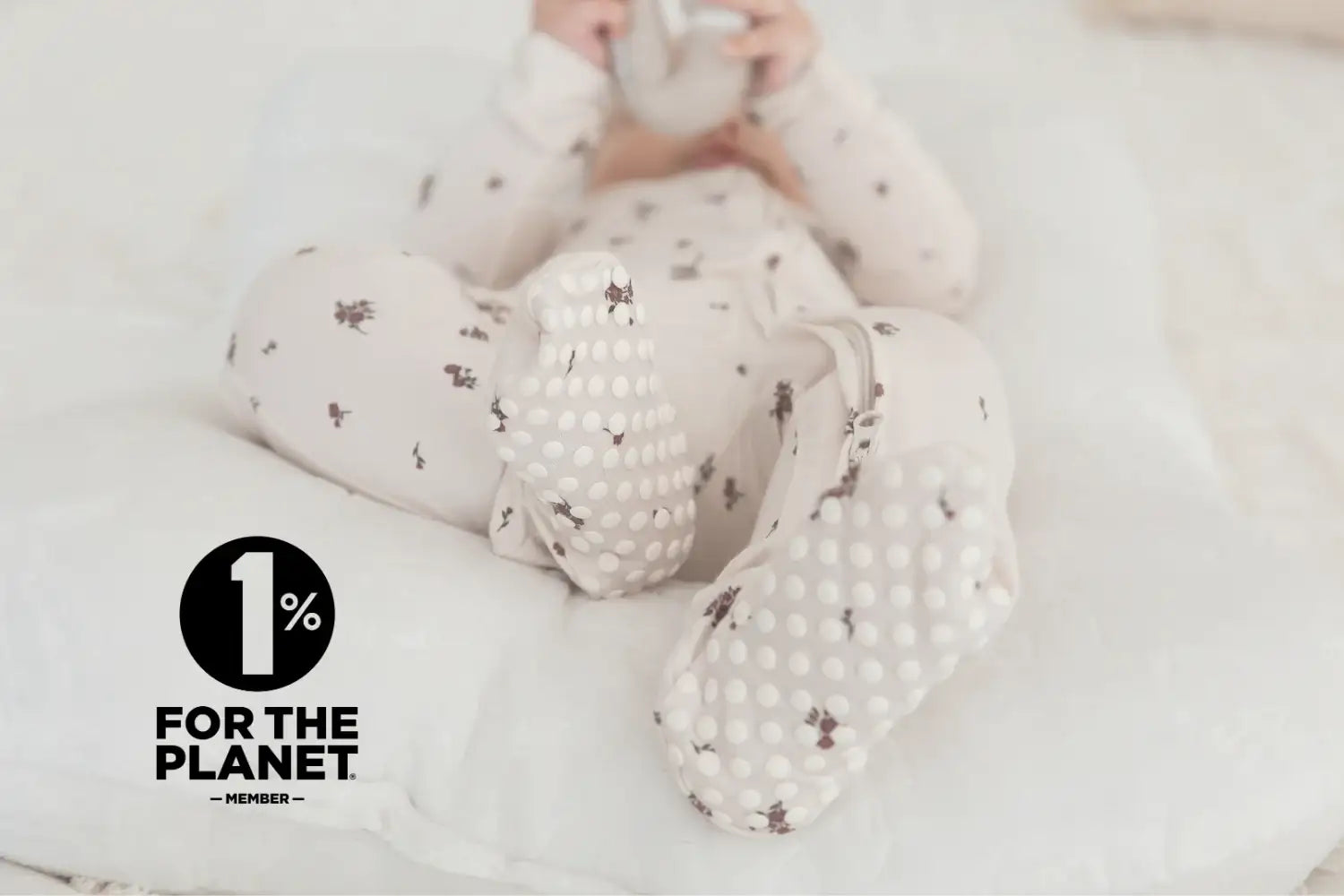A baby wearing the Milana Label Birch Floral footie, lying on a white quilted lounger. A black logo in the bottom-left corner reads '1% FOR THE PLANET MEMBER'.