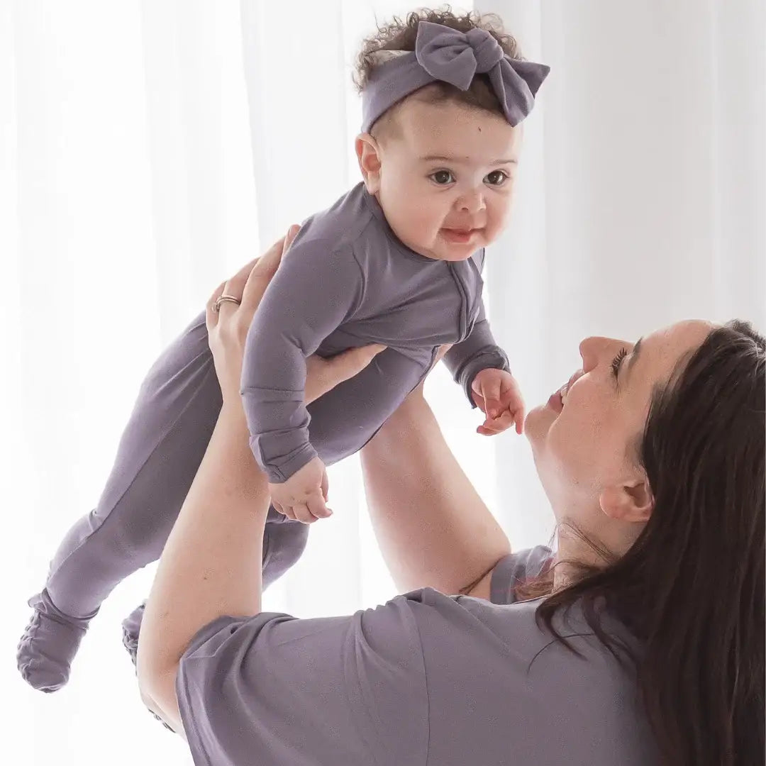A baby wearing a matching purple long-sleeve footed one-piece and headband with a large bow, being held up by a woman wearing a lounge set  in a bright, white-lit room.