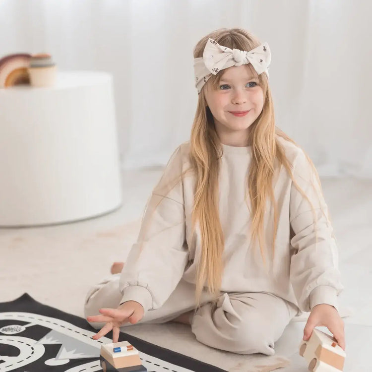 A child wearing a greige sweatshirt and pants set with a matching bow headband.