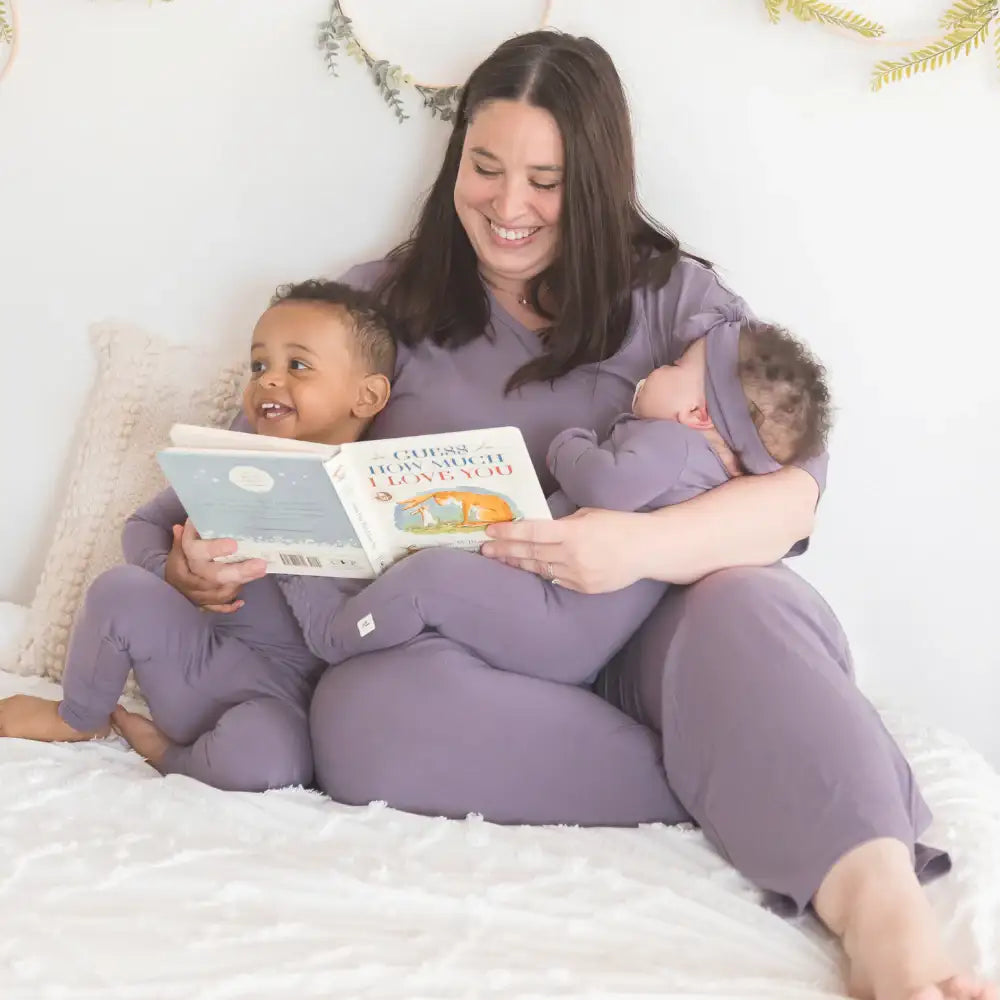 A mother wearing Milana Soft™ purple loungewear sits on a cozy bed reading a book to her toddler and holding her sleeping baby in matching purple bamboo TENCEL™ pajamas.