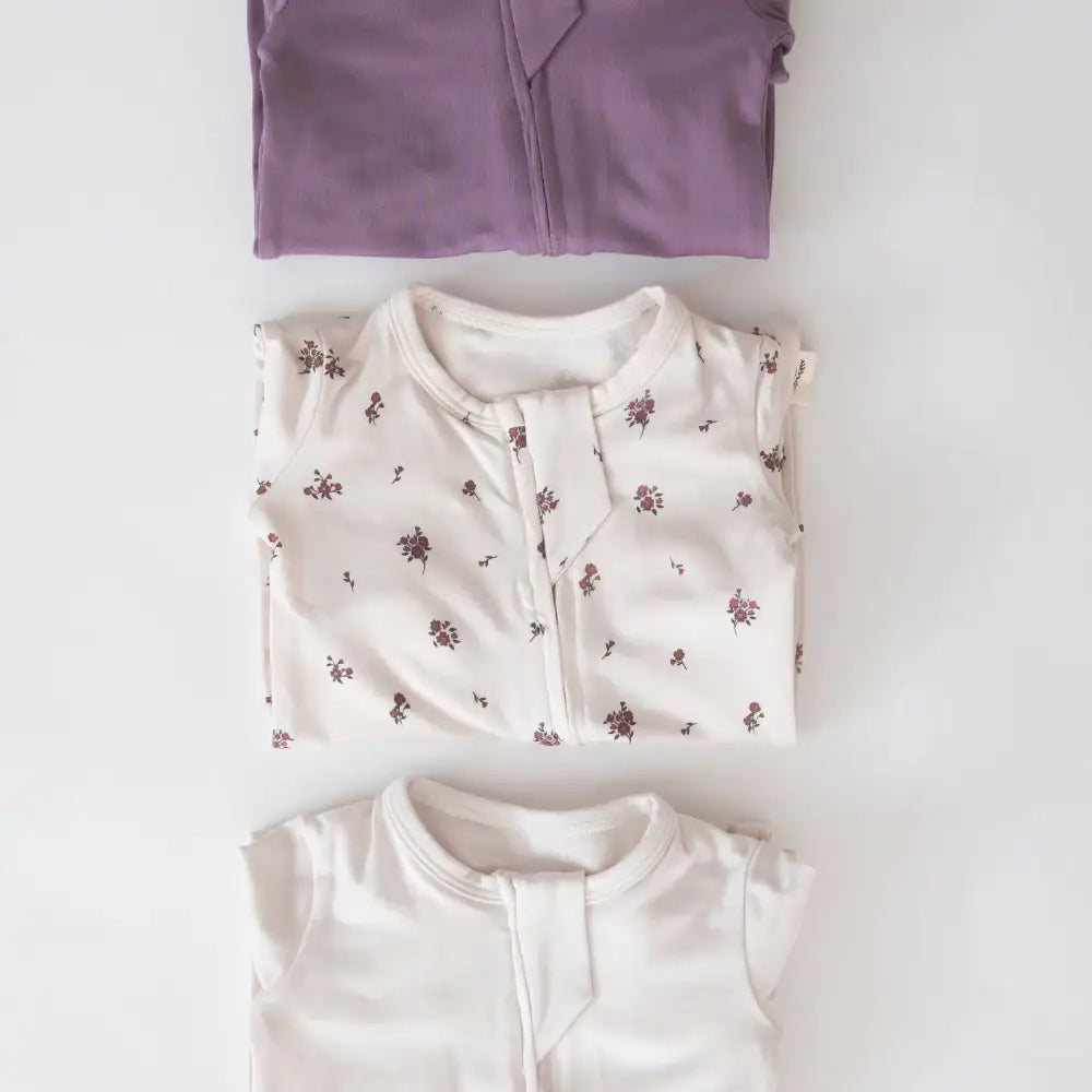 Three folded Milana Soft™ baby sleepers neatly stacked on a clean off-white background, showcasing the Wild Mulberry solid, Birch Floral pattern, and Birch solid fabric styles.