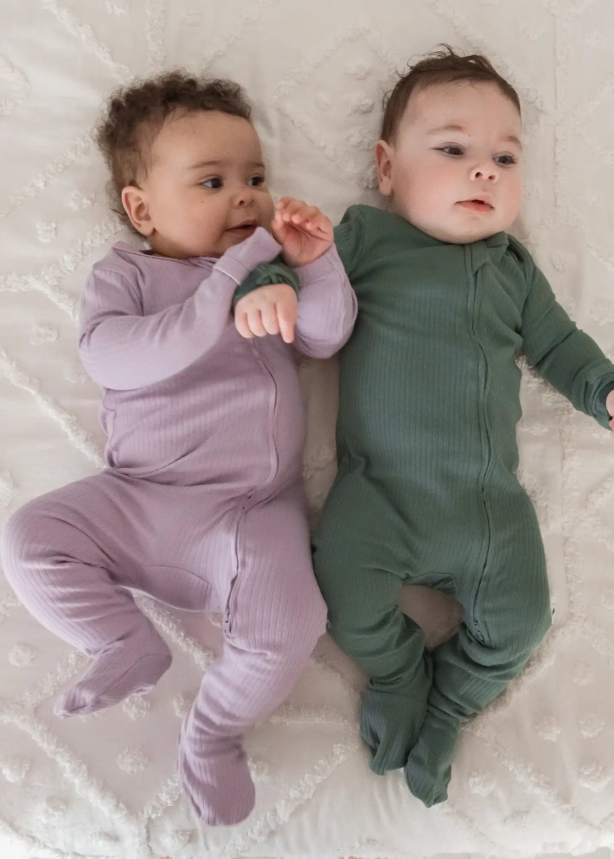 Two babies wearing matching ribbed zipper sleepers in the colours Lavender Blush and Pine.