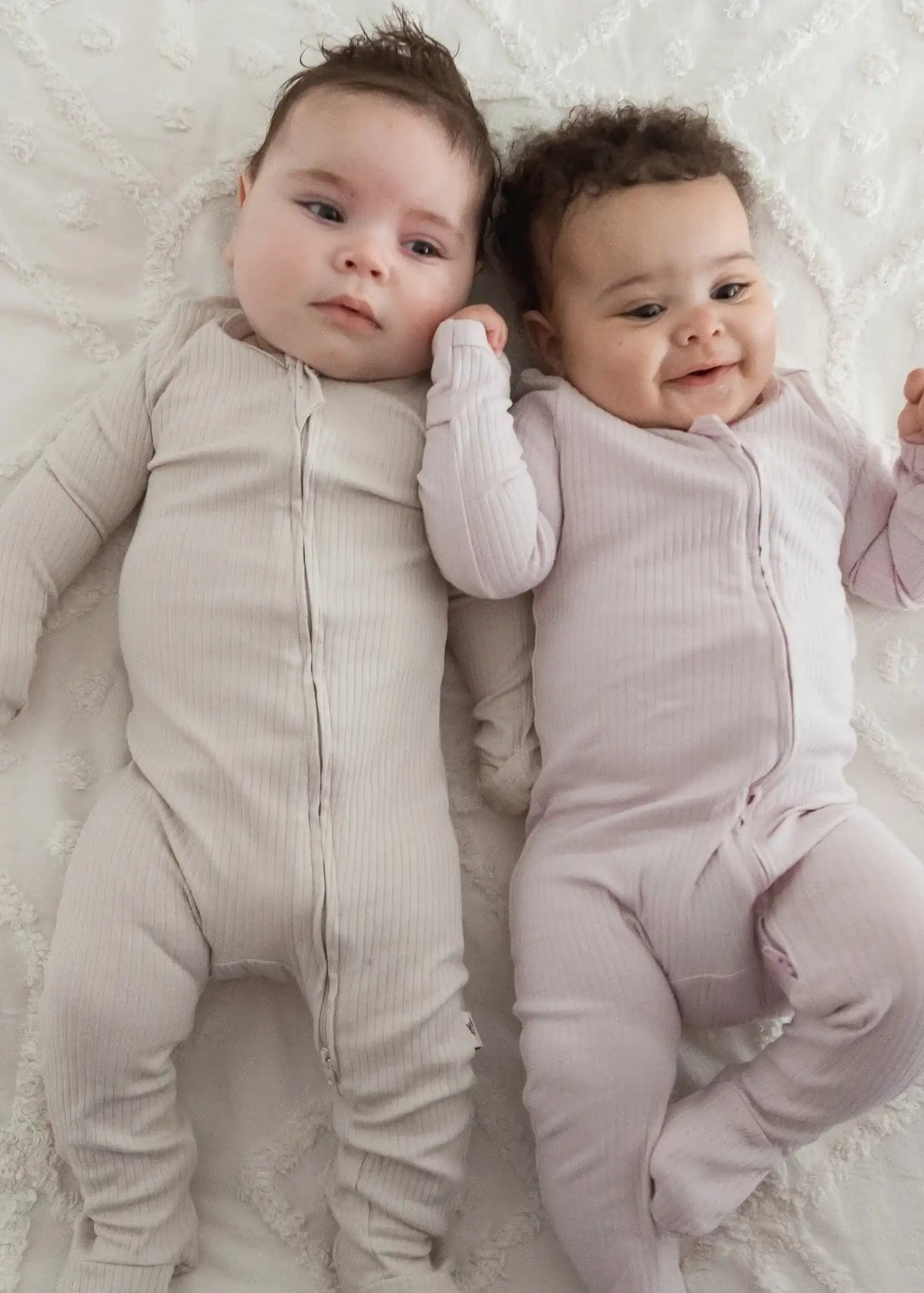 Two babies lying on a neutral blanket wearing a Milana sleeper in the colours Stone and Petal.