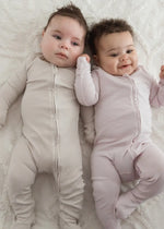 Two babies lying on a neutral blanket wearing a Milana sleeper in the colours Stone and Petal.