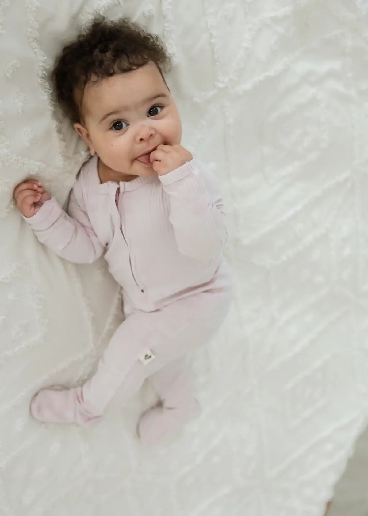 A baby lying on a neutral blanket in a Petal pink organic cotton footie PJ.