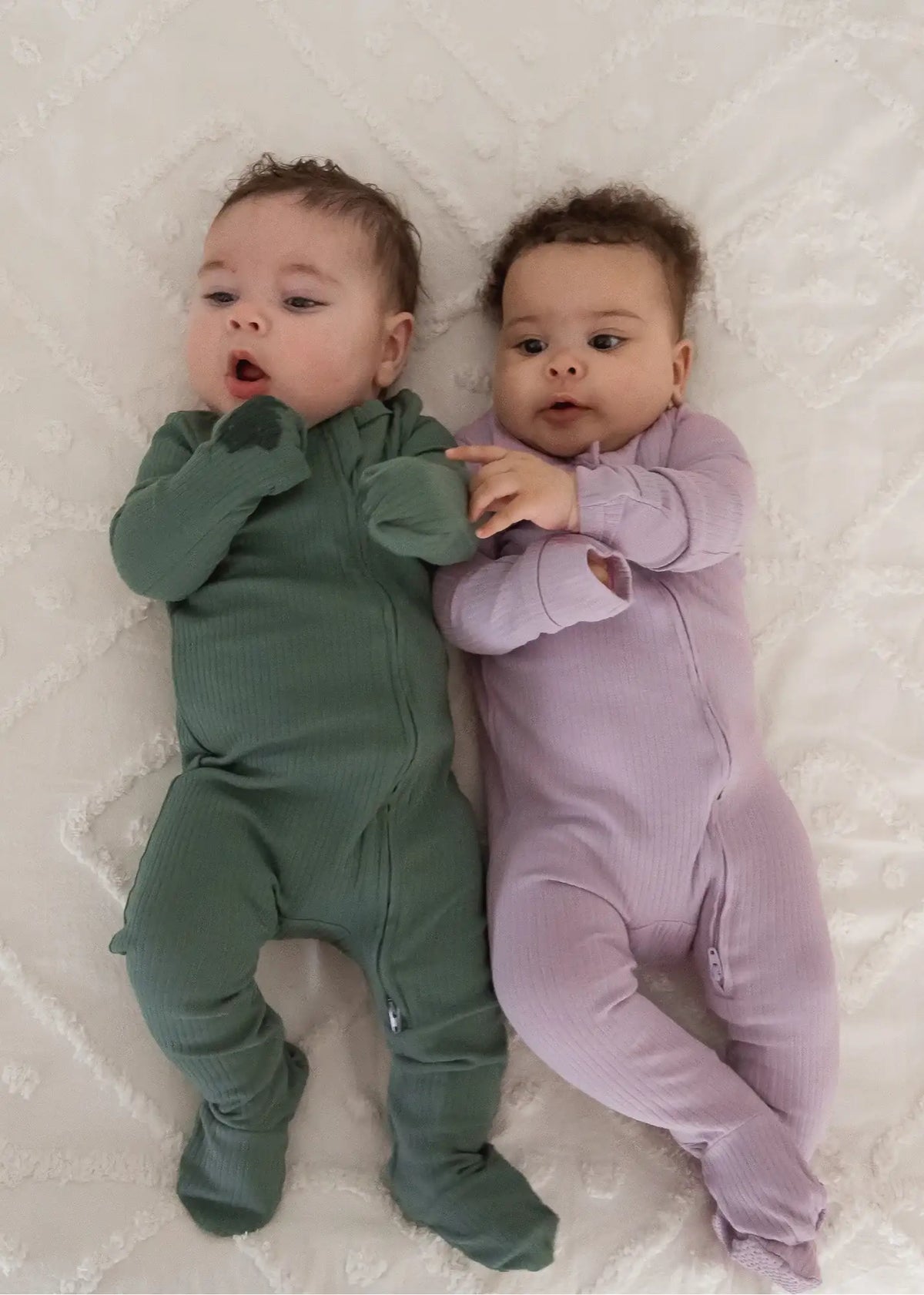 Two babies lying on a neutral blanket wearing ribbed sleepers in Pine and Lavender Blush.