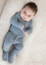 A smiling baby wearing a Soft Slate blue organic cotton sleeper.