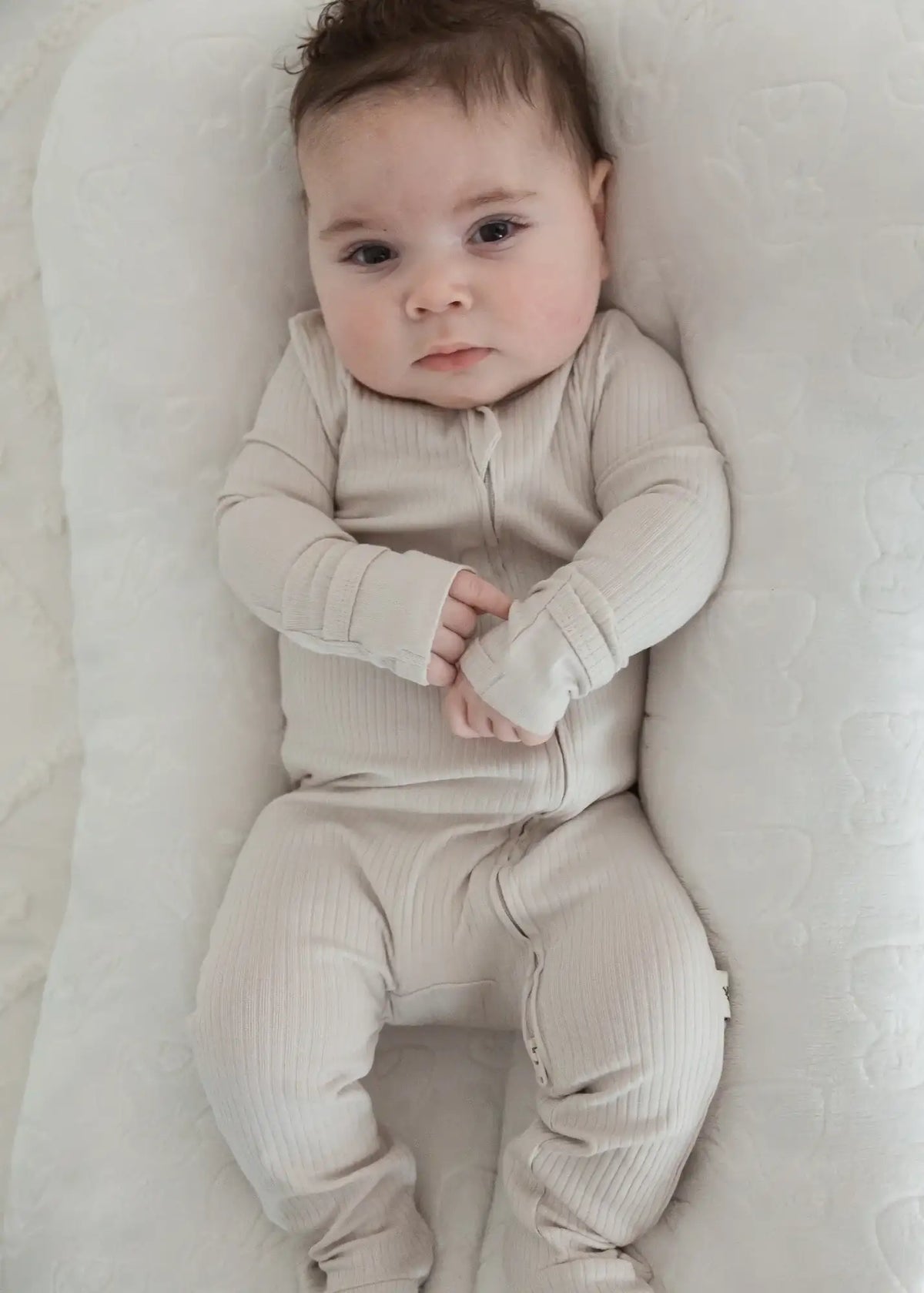 A baby lying in a neutral lounger while wearing an organic cotton sleeper in the colour Stone.