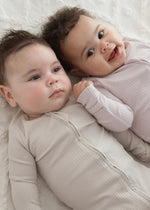 Two babies lying on a neutral blanket wearing matching zippered sleepers in the colours Stone and Petal.
