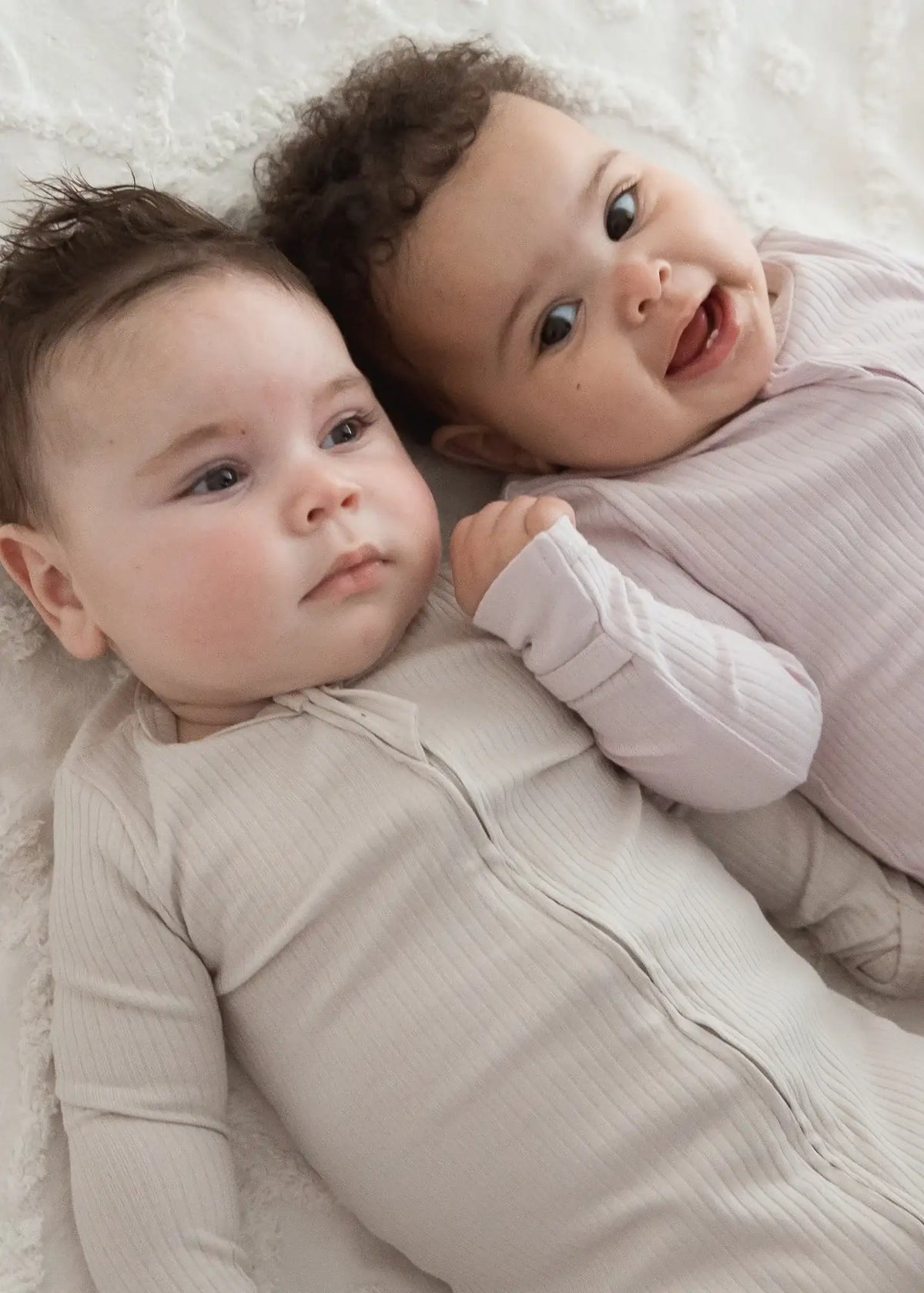 Two babies lying on a neutral blanket wearing matching zippered sleepers in the colours Stone and Petal.