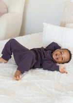 A toddler smiling at the camera while wearing a matching two-piece set in the colour Elderberry.