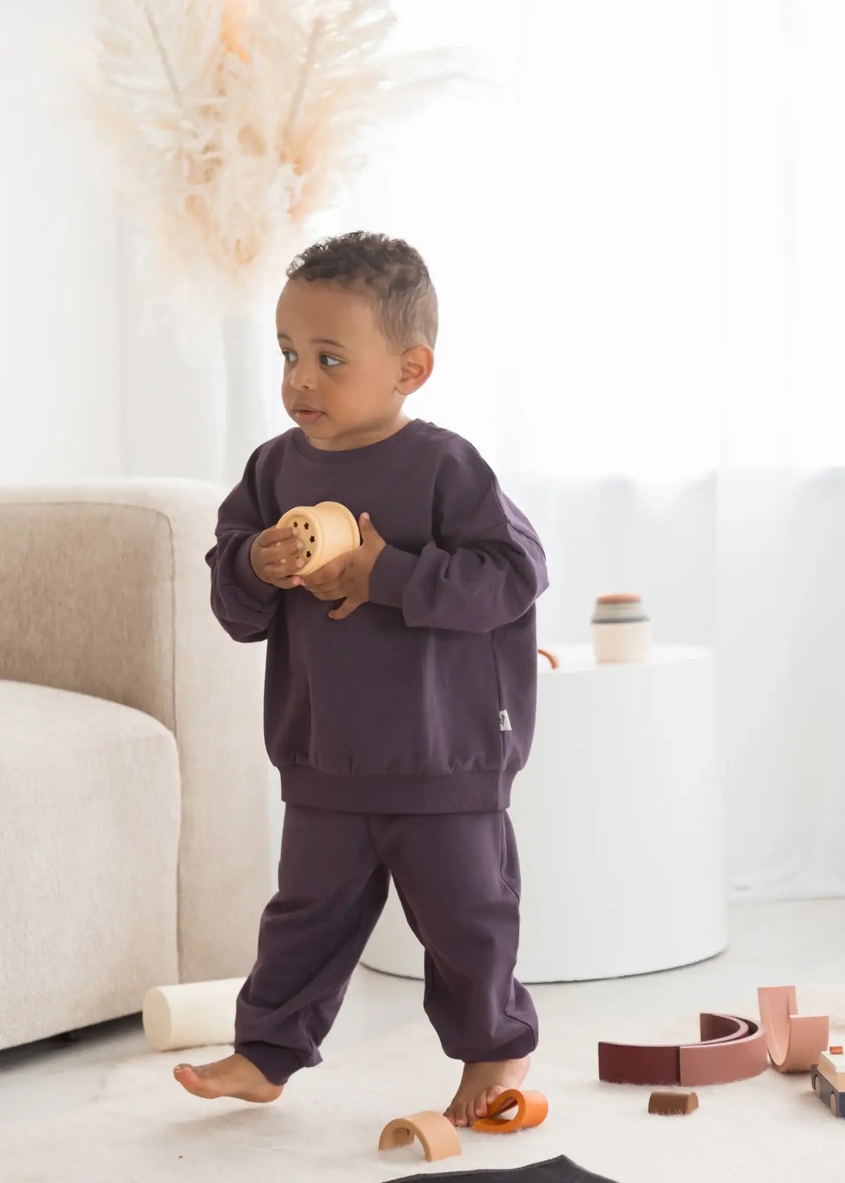A young boy walking in a bright room wearing an oversized sweatshirt and matching pants.