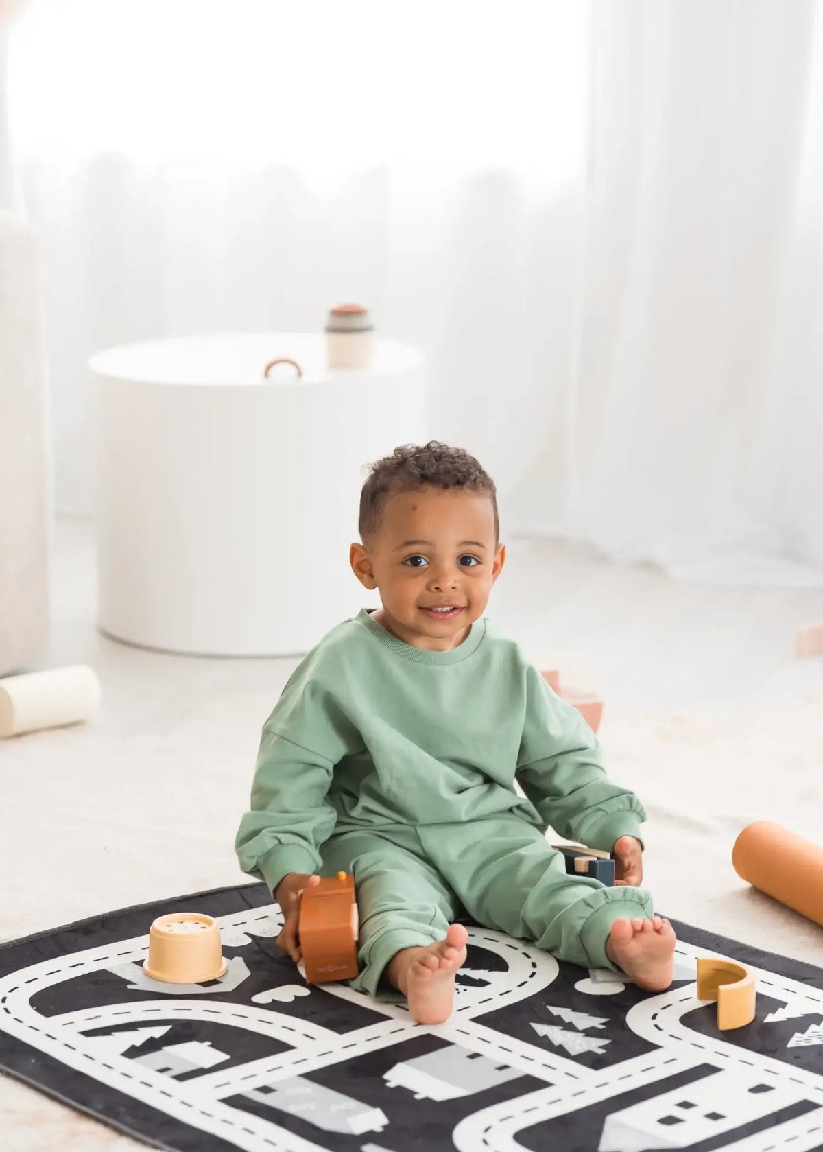 A toddler sitting on a play mat wearing an organic jogger set in the colour Eucalyptus green.