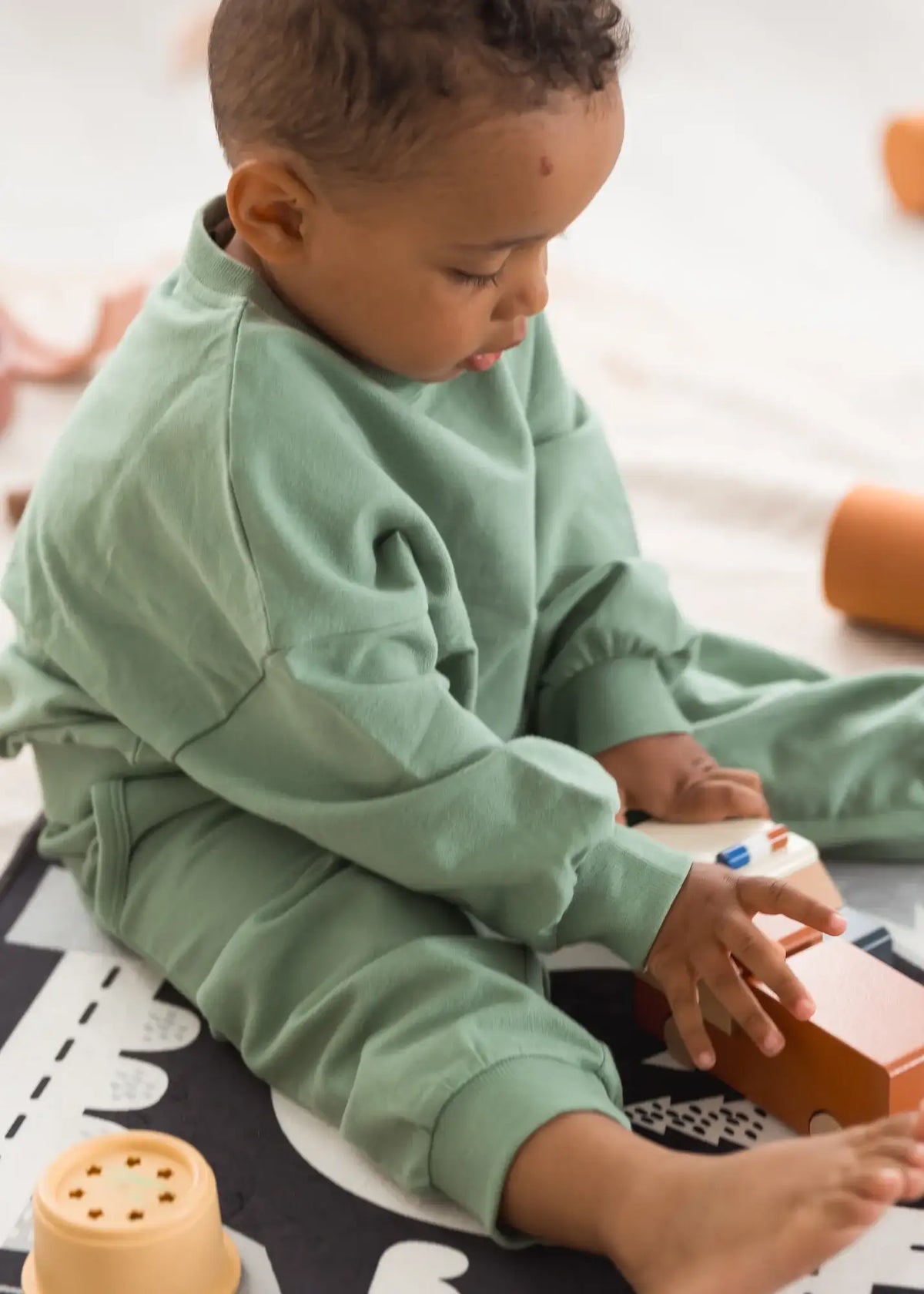 A toddler sitting on the floor playing with wooden cars in a Eucalyptus green jogger set.
