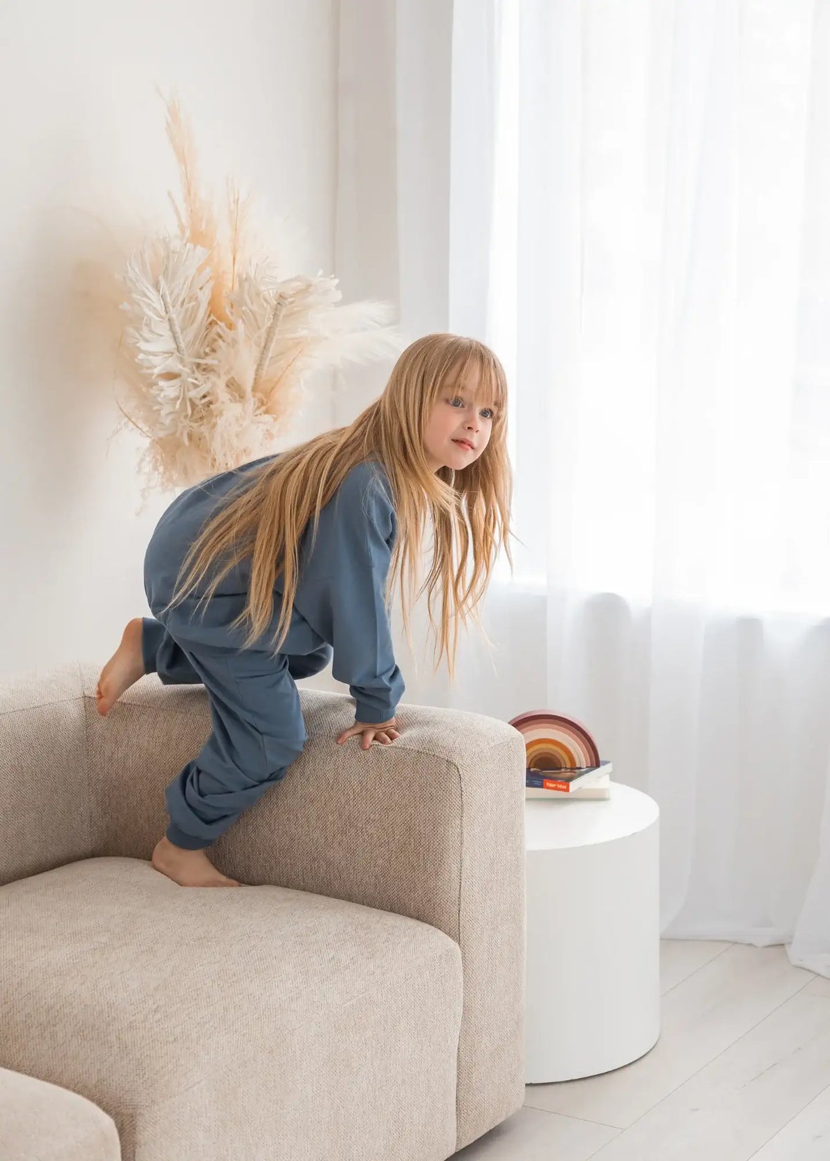 A young girl climbing on a neutral sofa wearing an oversized jogger set by Milana Label.
